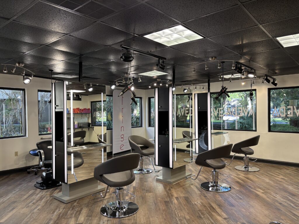 Modern hair salon interior with multiple black styling chairs, mirrors, wood flooring, and large windows letting in natural light. - Fringe Salon, Hiltonhead, SC