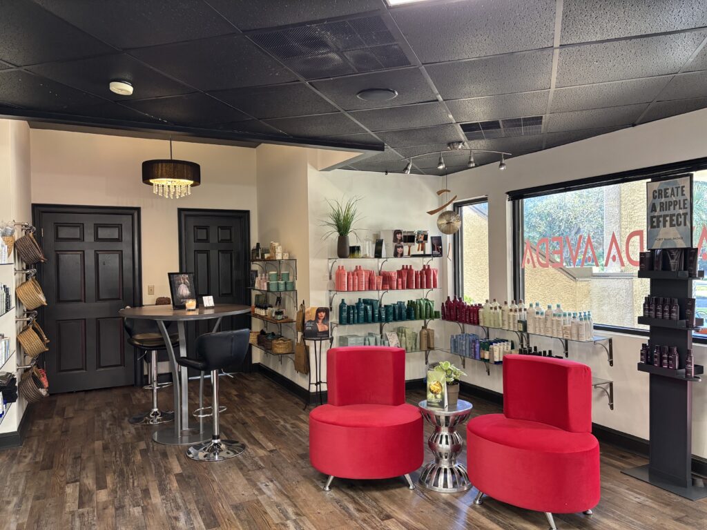 Modern salon interior with two red chairs, shelves stocked with hair products, a round table with stools, and large windows allowing natural light. - Fringe Salon, Hiltonhead, SC