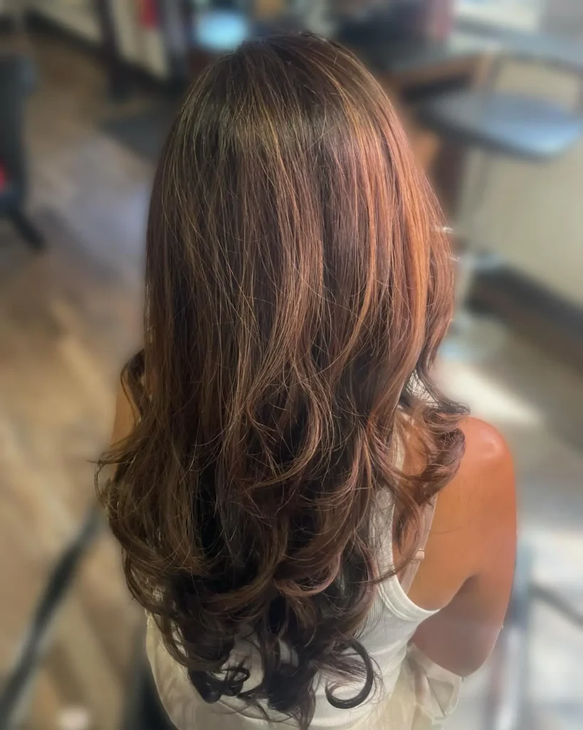 A person with long, wavy, brown hair sits in a salon chair, facing away from the camera. - Fringe Salon, Hiltonhead, SC