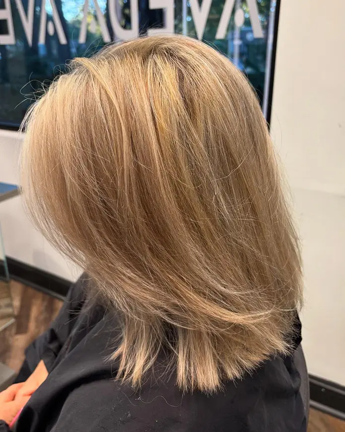 A person with straight, shoulder-length blonde hair sits in a salon chair, wearing a black cape, with salon signage visible in the background. - Fringe Salon, Hiltonhead, SC