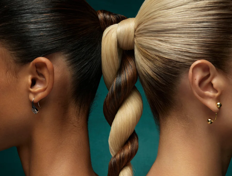 Two women stand back to back with their hair, one dark and one blonde, braided together in a single twisted ponytail; both wear earrings. - Fringe Salon, Hiltonhead, SC
