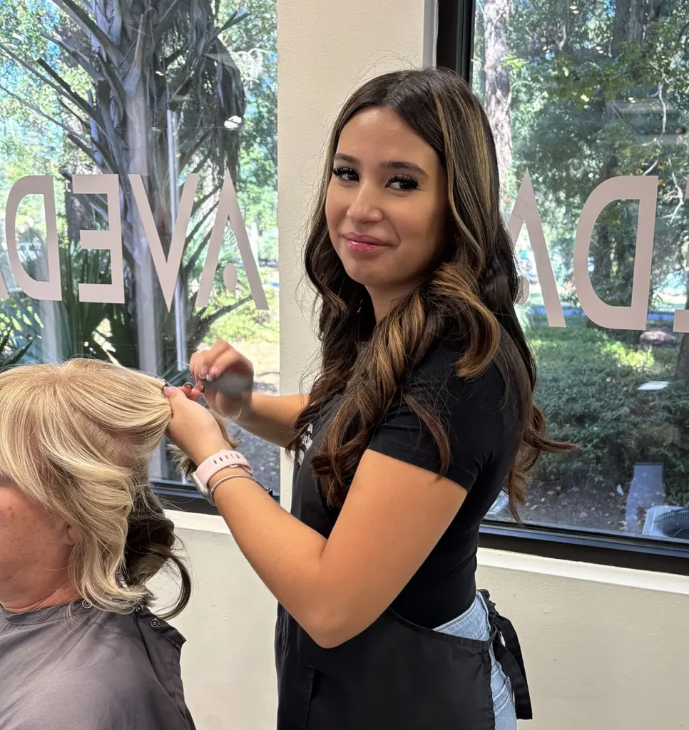 A young woman styles another person's hair in a salon, standing by a window with trees visible outside. - Fringe Salon, Hiltonhead, SC