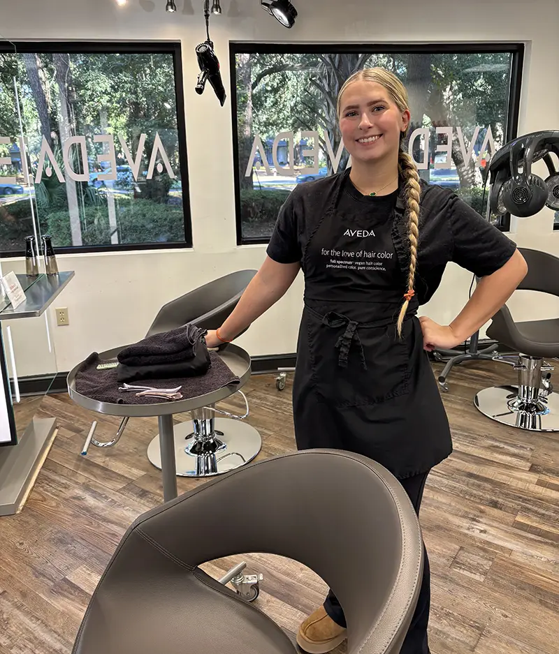 A hairstylist with a long braid stands smiling beside a salon chair, wearing an Aveda apron in a modern, well-lit salon. - Fringe Salon, Hiltonhead, SC