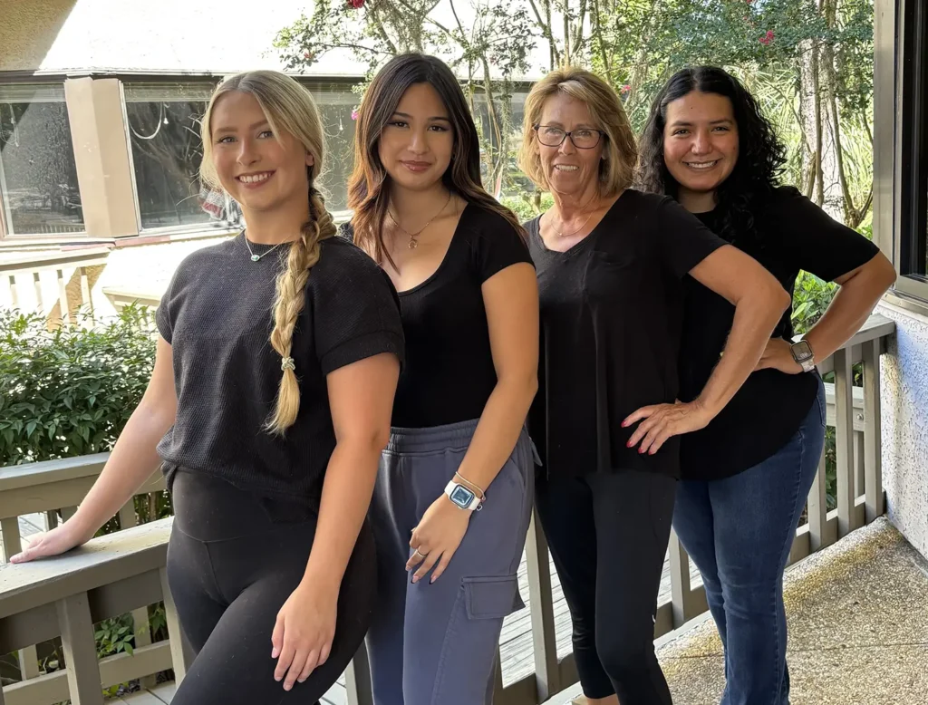 Four women stand side by side on a porch, smiling at the camera. They are casually dressed in black tops, with greenery and a house visible in the background. - Fringe Salon, Hiltonhead, SC