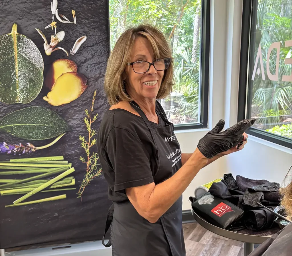 A woman wearing glasses, a black t-shirt, and black gloves stands in a salon, preparing hair supplies near a window and a poster with botanical images. - Fringe Salon, Hiltonhead, SC