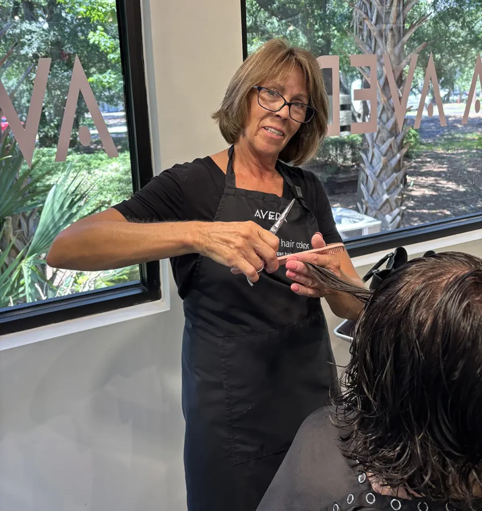 A hairstylist wearing an apron is cutting the hair of a seated client inside a salon with large windows. - Fringe Salon, Hiltonhead, SC