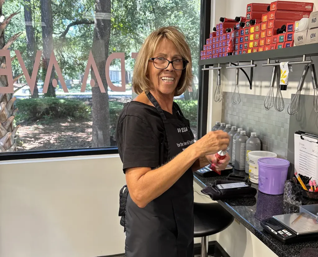 A woman in a black apron stands at a salon workstation, holding a hair product bottle and smiling at the camera. Shelves hold hair color boxes and supplies. - Fringe Salon, Hiltonhead, SC