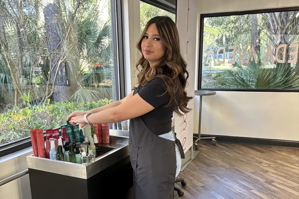 A woman in a salon apron stands beside a cart with hair products, looking toward the camera. Large windows show trees and greenery outside. - Fringe Salon, Hiltonhead, SC