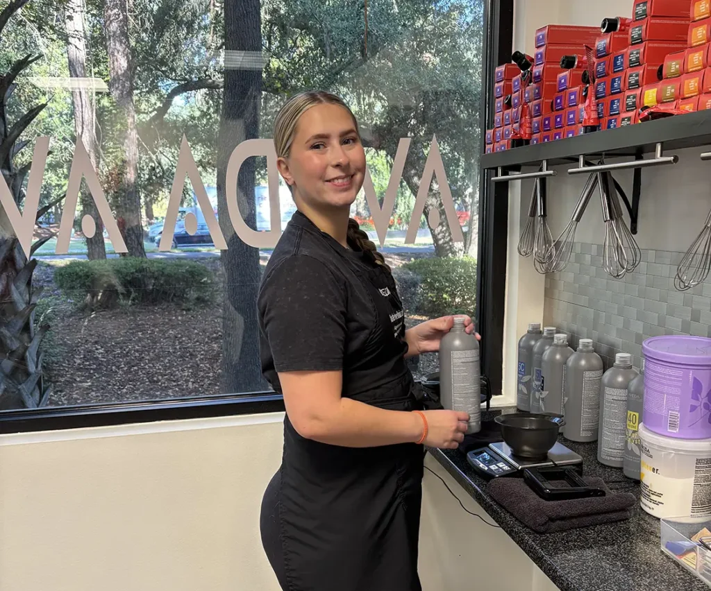 A person wearing a black apron stands at a counter with hair products, smiling at the camera inside a salon with a window showing trees outside. - Fringe Salon, Hiltonhead, SC