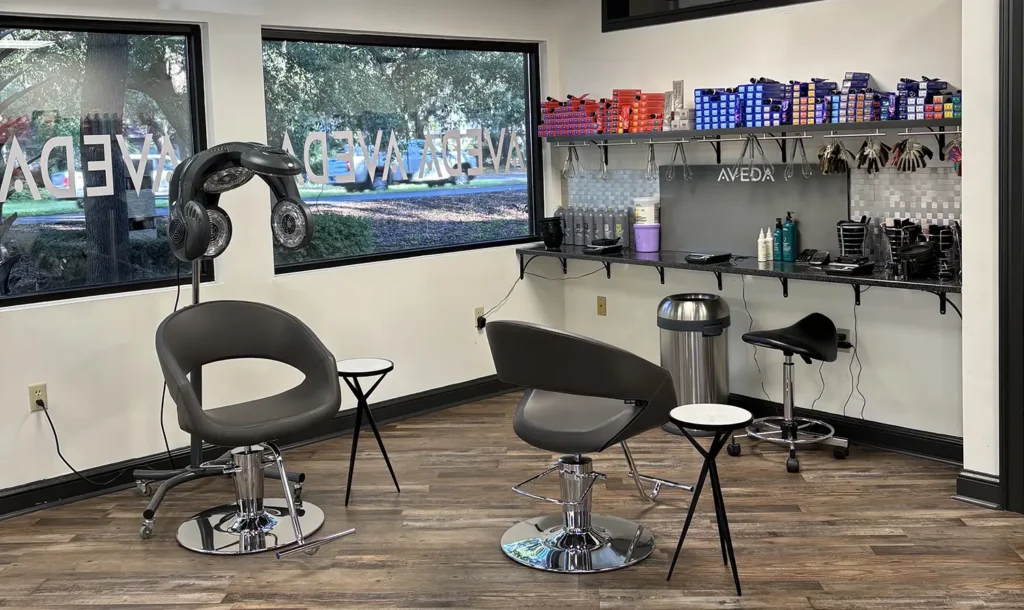 A modern hair salon with two styling chairs, hair dryers, a work counter with hair products, tools, a stool, and large windows overlooking trees. - Fringe Salon, Hiltonhead, SC